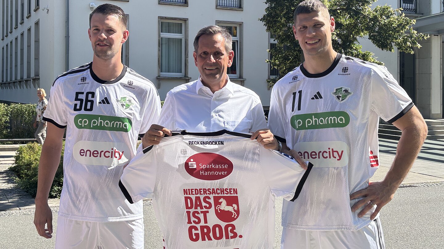 Ministerpräsident Olaf Lies hält ein Handballtrikot mit dem Slogan „Niedersachsen. Das ist groß." Zwei Spieler des TSV Hannover-Burgdorf stehen neben ihm.