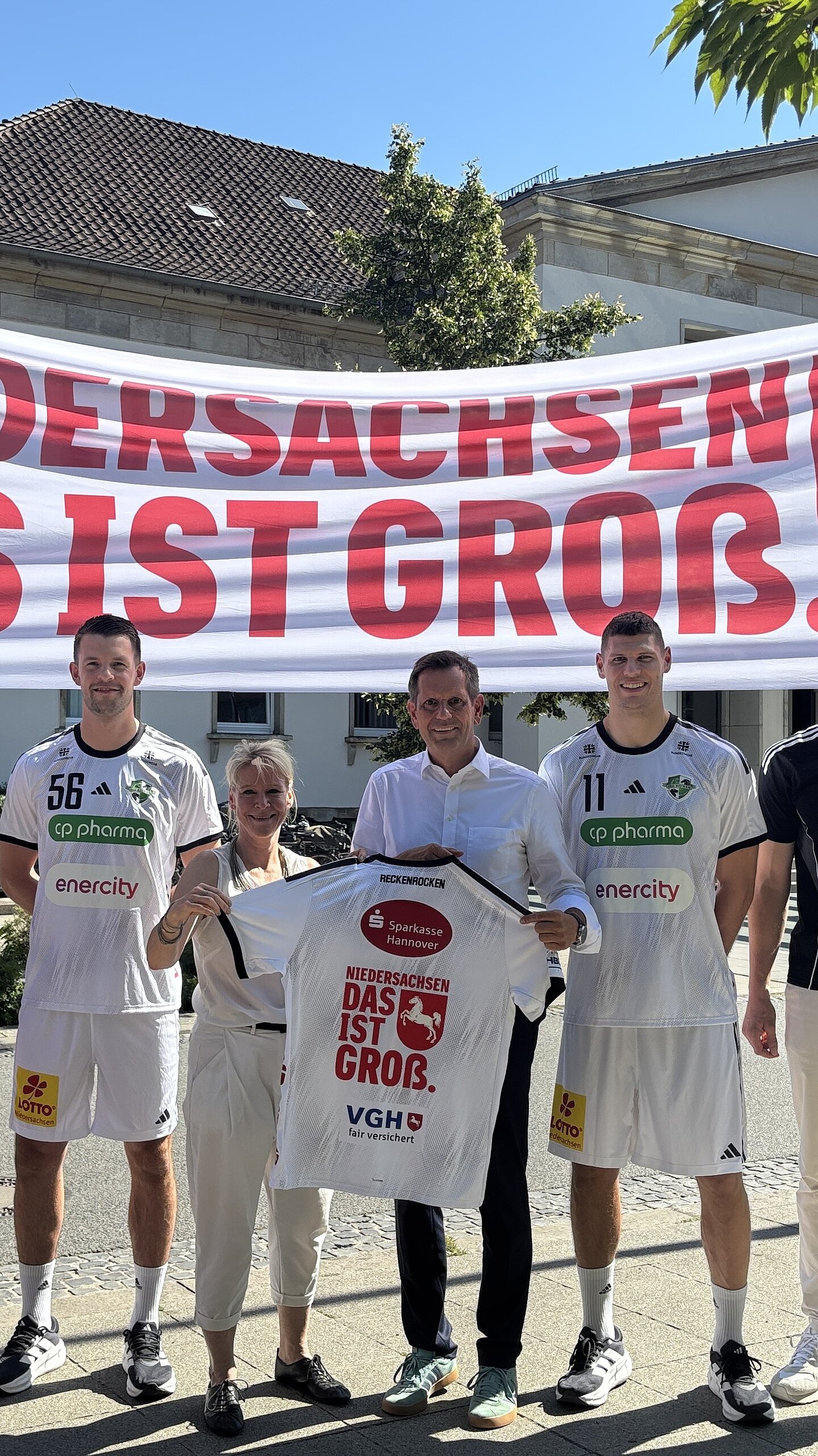 Gruppenfoto bei Sonnenschein. Zwei Männer halten ein großes Banner mit der Aufschrift „Niedersachsen. Das ist groß." und dem Sachsenross. Ein Mann hält ein Handballtrikot.