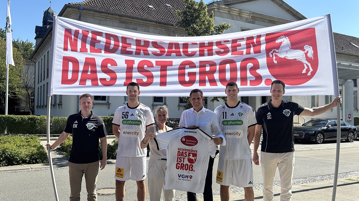 Gruppenfoto bei Sonnenschein. Zwei Männer halten ein großes Banner mit der Aufschrift „Niedersachsen. Das ist groß." und dem Sachsenross. Ein Mann hält ein Handballtrikot.